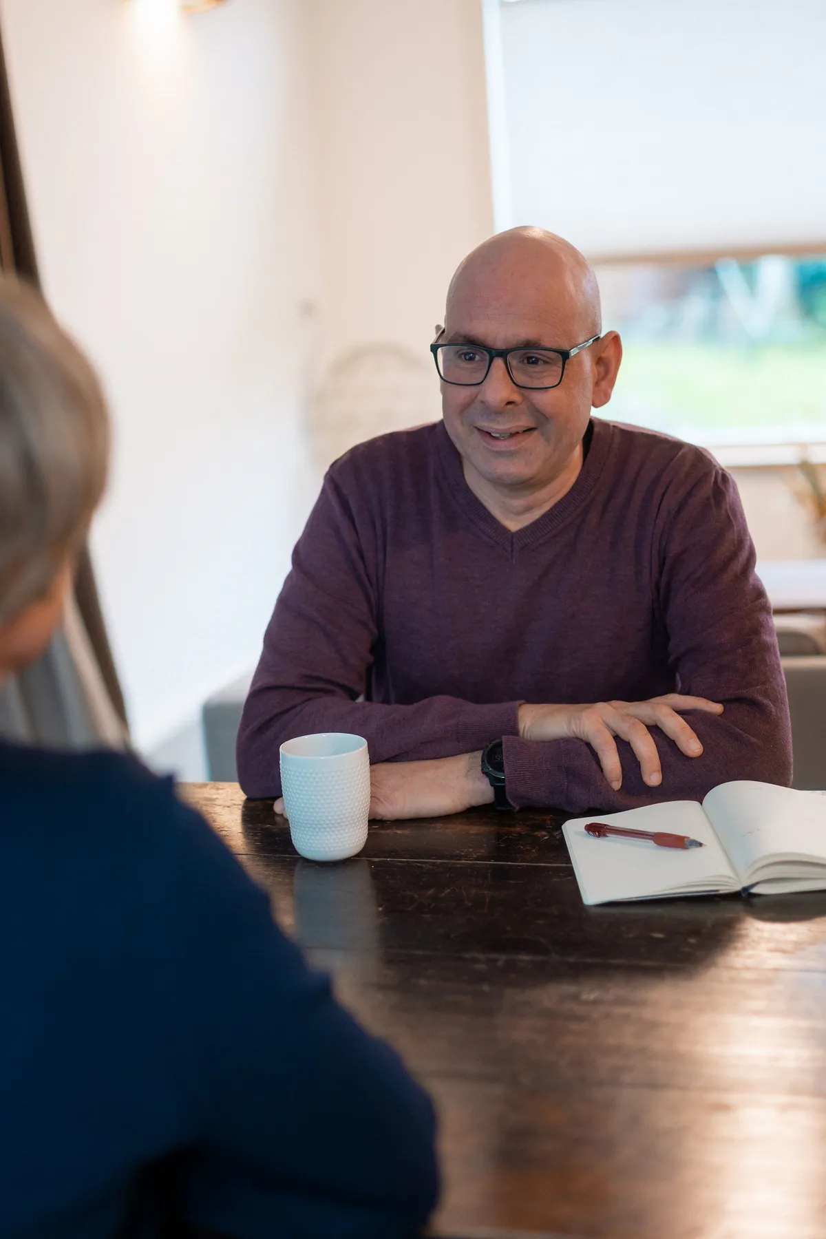 Roeland in gesprek aan een houten tafel, met een mok koffie en een notitieboek.